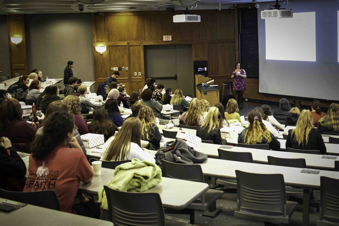 A group of attendees sits in a large lecture hall to watch a presentation.