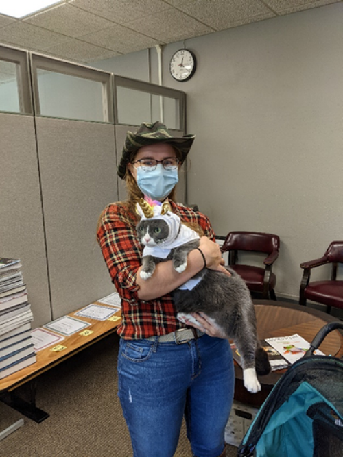 A photo of Sarah Stangl wearing a plaid shirt, jeans, and holding her cat, who is wearing a unicorn outfit.