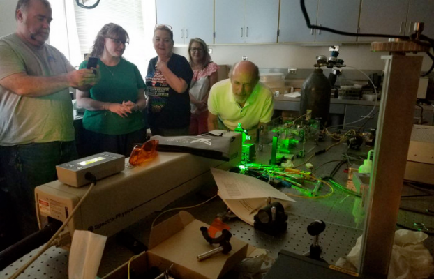 A group of people gather in a dark room to observe an experiment set up on a lab table.