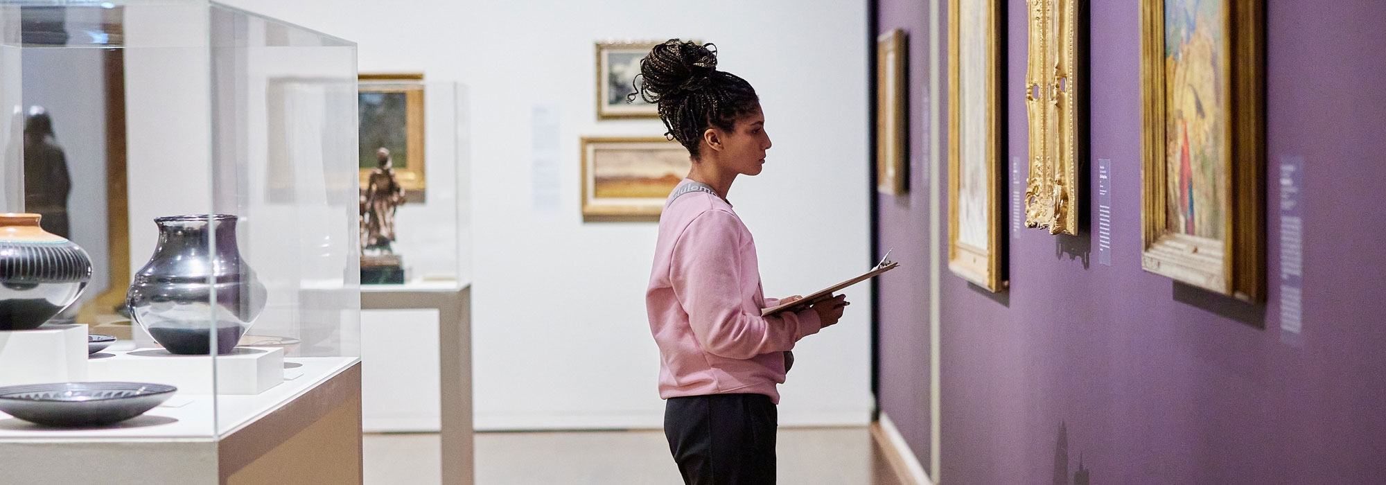 A student holding a clipboard stands in front of a historical painting in a museum. Other artifacts and paintings are nearby.