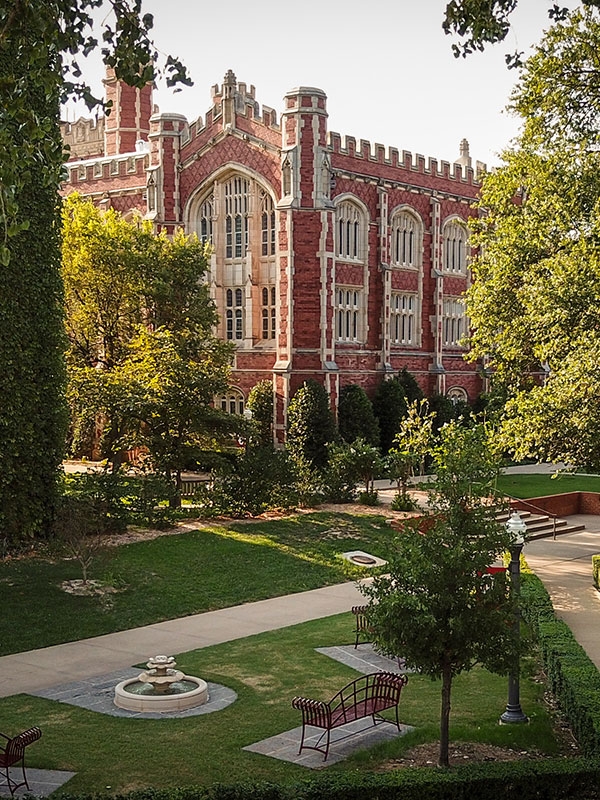 A view of Evans Hall highlighting a sidewalk lined with green trees, a bench, and fountain.