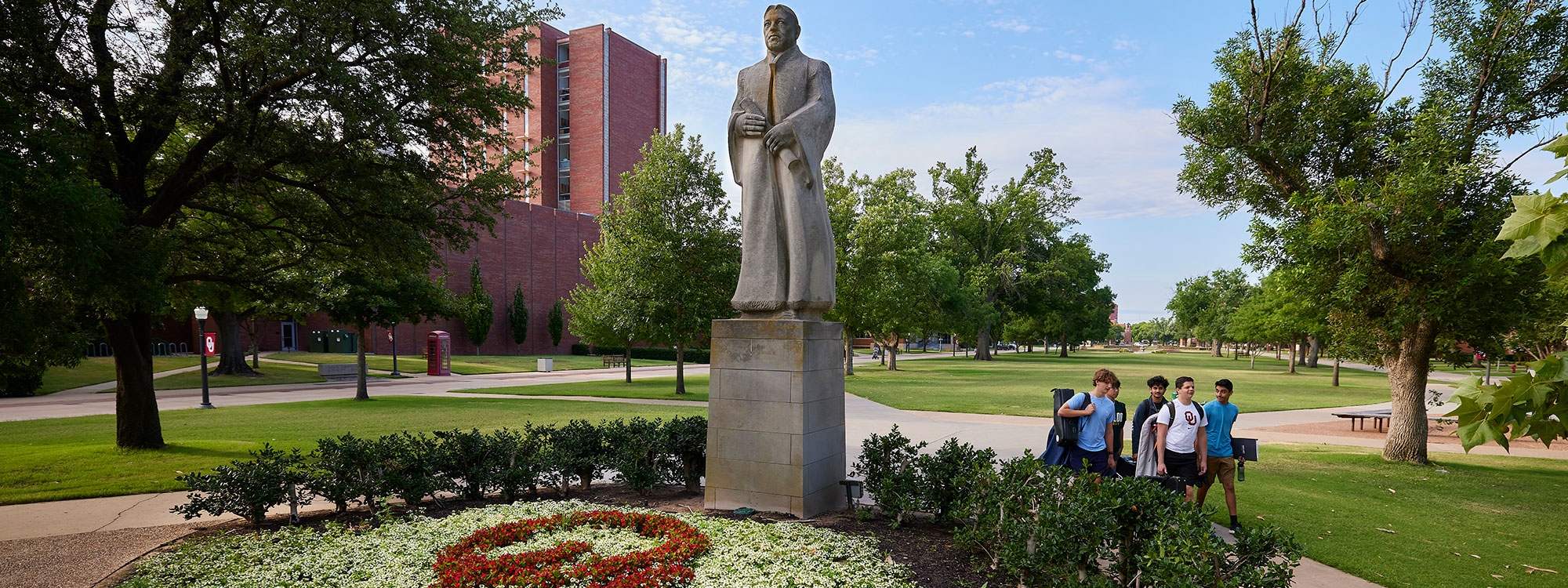 A view of summer on the South oval with blooming flowers and students walking pass a large concrete statue of Bizzell.