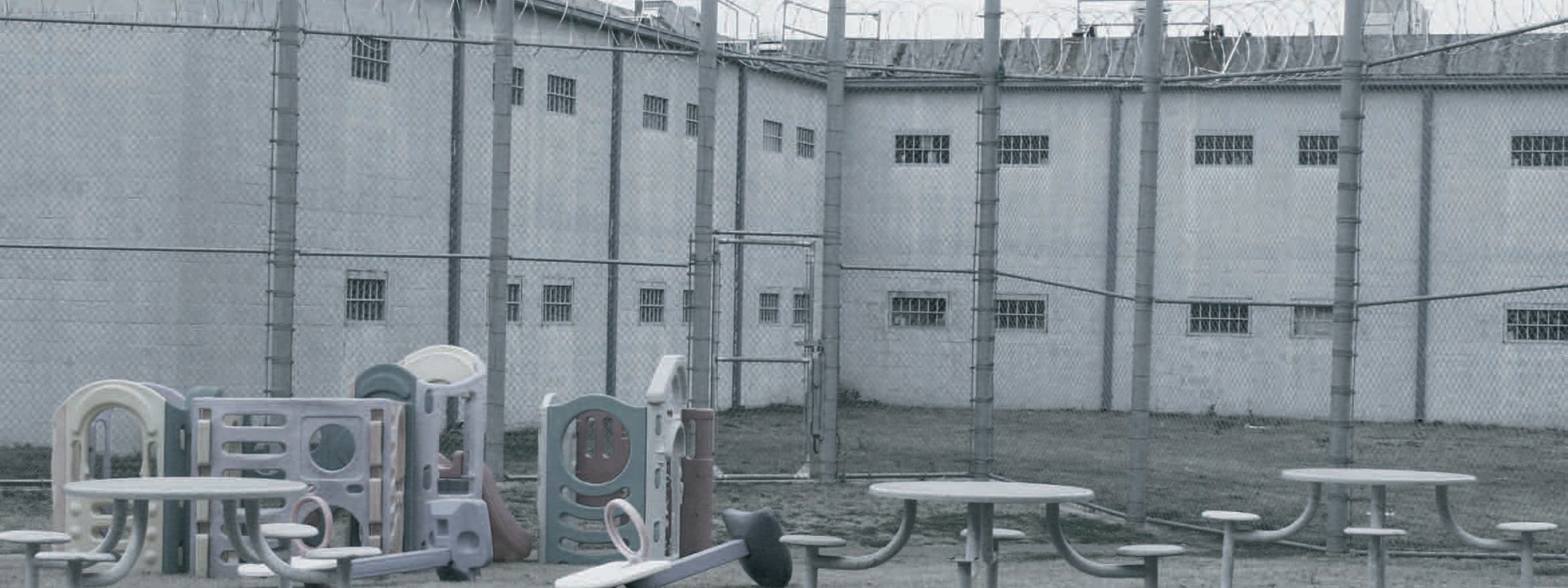 A prison, with a barbed-wire fence, picnic tables, and playground equipment in the foreground.
