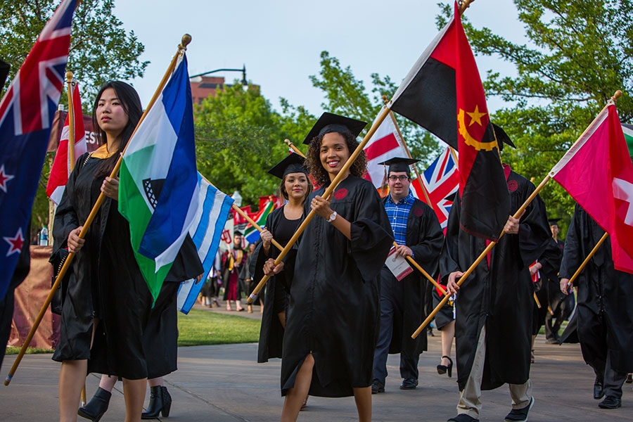 International students in graduation regalia carrying flags in a parade.