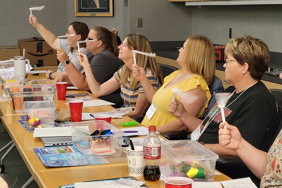Five teachers sitting at a long table slinging a scientific instrument to measure humidity in the air.