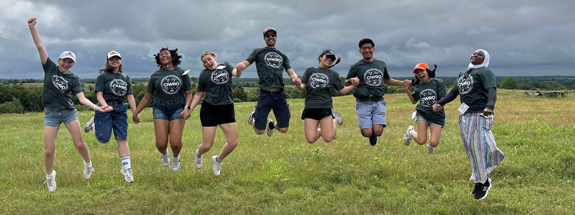 Students wearing green CIWRO t-shirts jump while holding hands on a grassy hill with cloudy skies in the background.