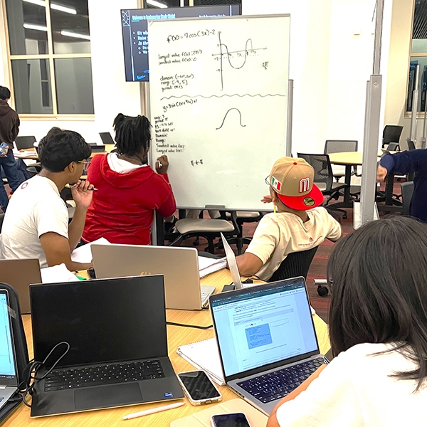 Students working in a large study group with laptops and whiteboards.