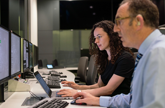 A student and a teacher sitting at a laptop and multiple computer monitors.