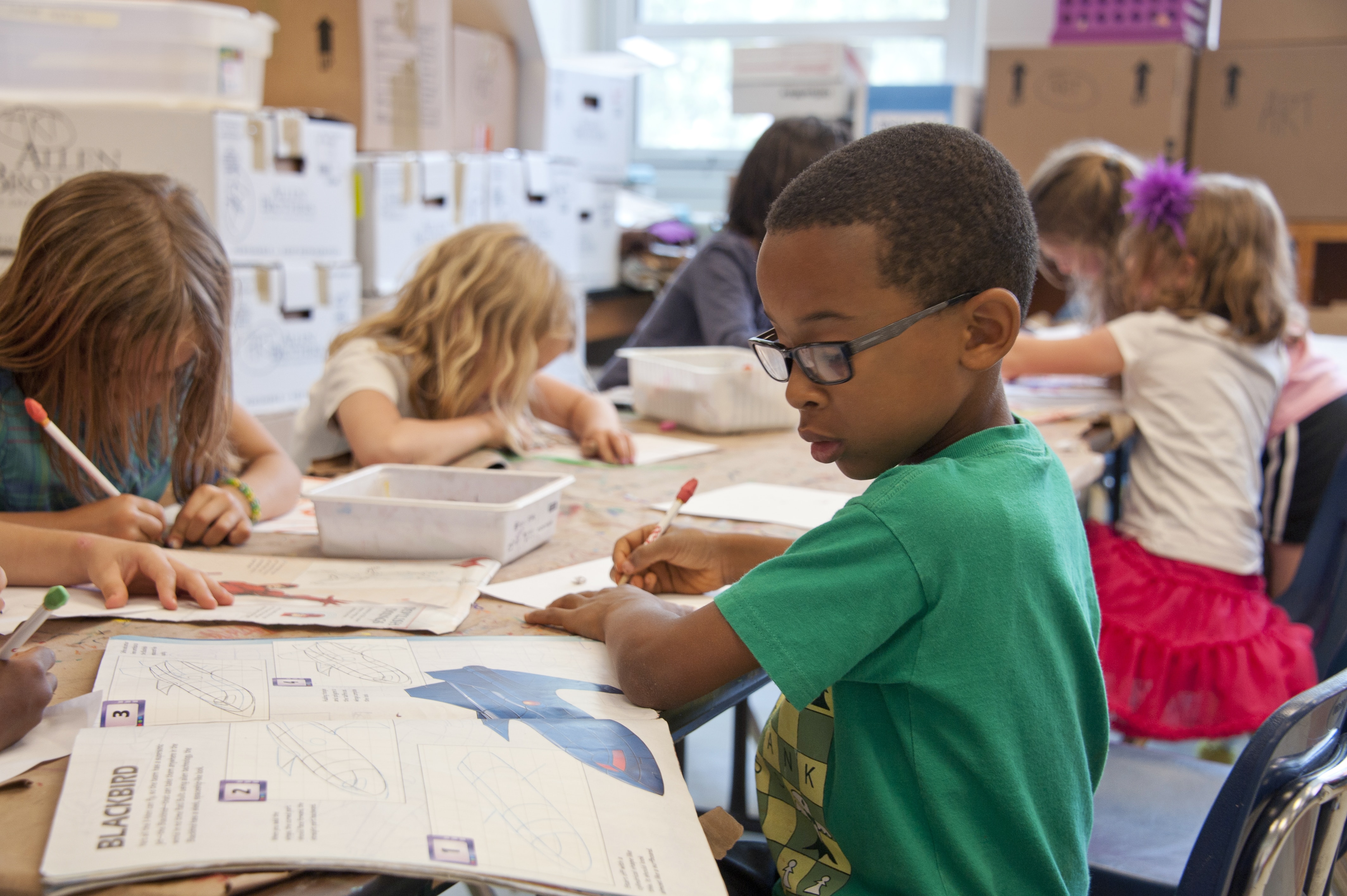 Stock image: a child works on a worksheet at a table with other children in a classroom setting.