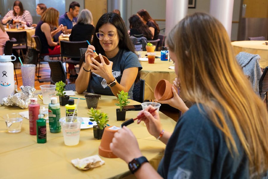 Two students painting flower pots with more students in the background. One student is facing the camera and the other is looking away. 