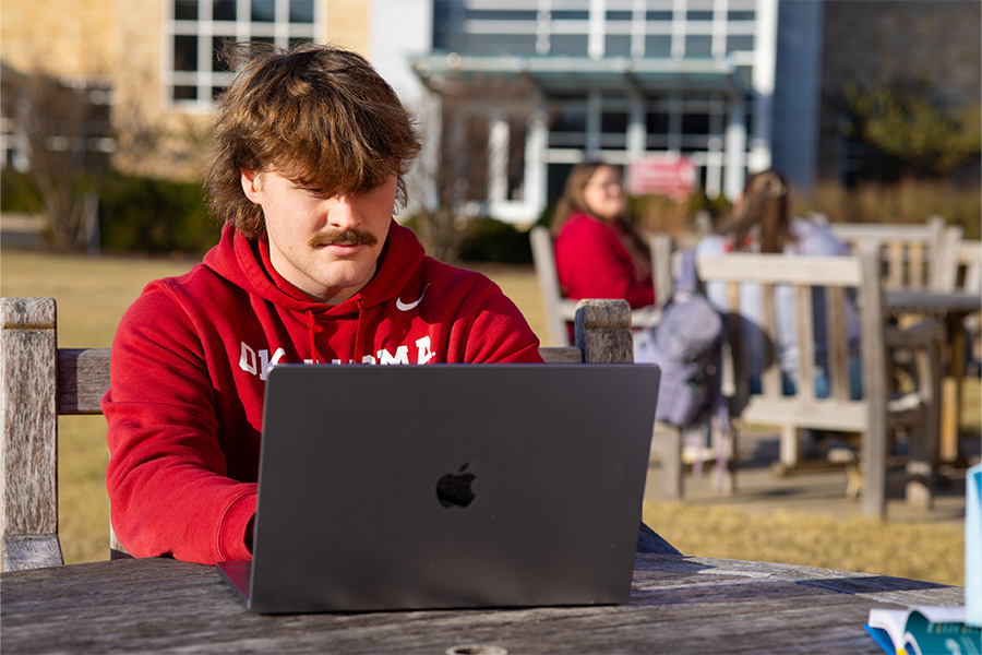 OU-Tulsa student sits outside and looks at a laptop screen.