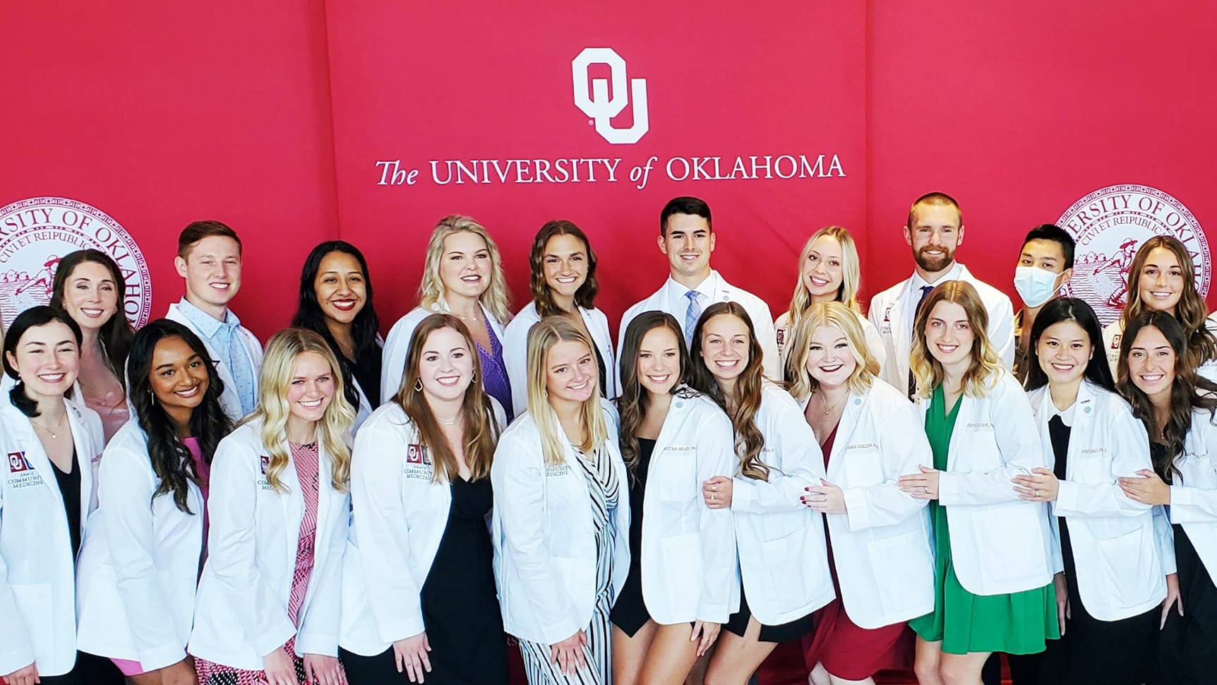 OU-Tulsa PA students in white coats stand in front of a red background with an OU logo on it