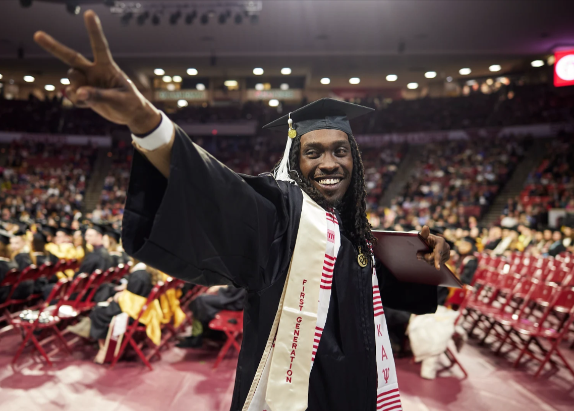 A student holding up a peace sign while walking in his cap and gown at graduation. 