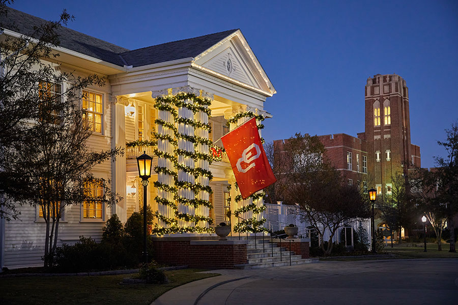 Boyd House at night, with lights and garland wrapped around columns and an OU flag hanging from the front.