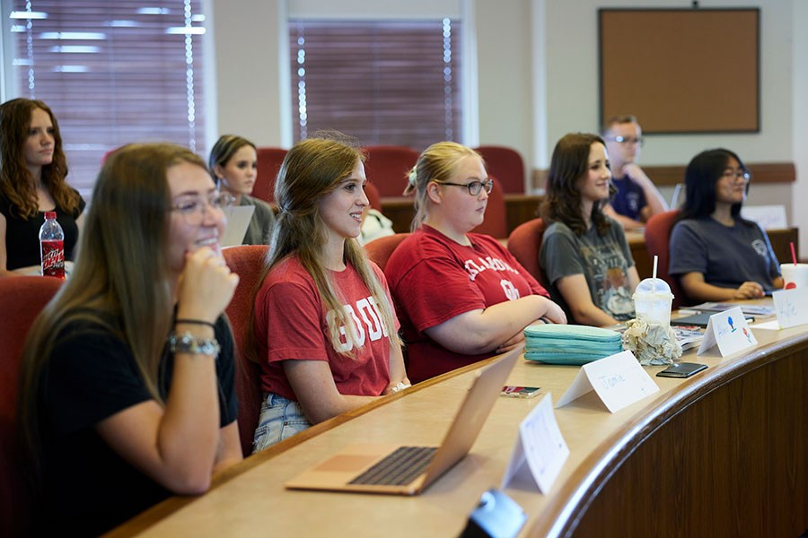 A classroom with rows of desks, where students are seated, each with name placards in front of them. Some desks have laptops and pencil cases. The students are smiling and looking toward the front of the classroom, attentively listening to the lecture or presentation.