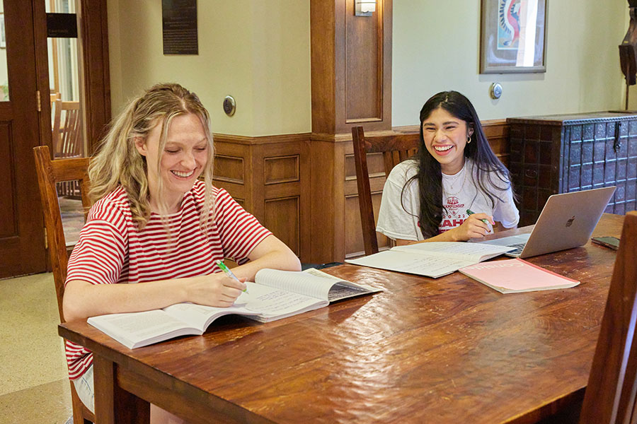 Two students sit at a desk in the Wagner Hall lobby, both smiling and writing in their notebooks. The student on the left has two books or notebooks on the desk and is writing while smiling, engaged in their work. The student on the right is also writing in their notebook, smiling at the student next to them.