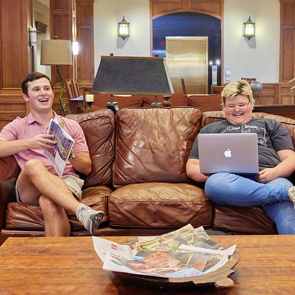 Two students sit at opposite ends of a couch in the Wagner Hall lobby, both smiling and relaxed. The student on the left is holding a school newspaper with their legs crossed, while the student on the right has a laptop resting on their crossed leg. The scene captures a casual, friendly moment in a common area, with both students engaged in their activities.