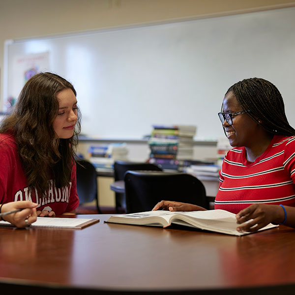 Two students sit together at a table in the learning center, engaged in conversation. The student on the left holds a pencil and has a notebook beside her, appearing ready to take notes. The student on the right has an open book in front of her and is smiling warmly as they talk. Both seem relaxed and engaged in their discussion.