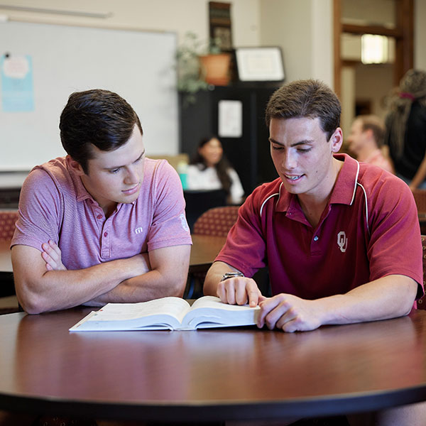 Two students sit side by side at a table, both focused on a book they are sharing. The student on the left has their arms crossed, listening attentively to the other student. The student on the right is pointing to a specific line in the book while explaining something.