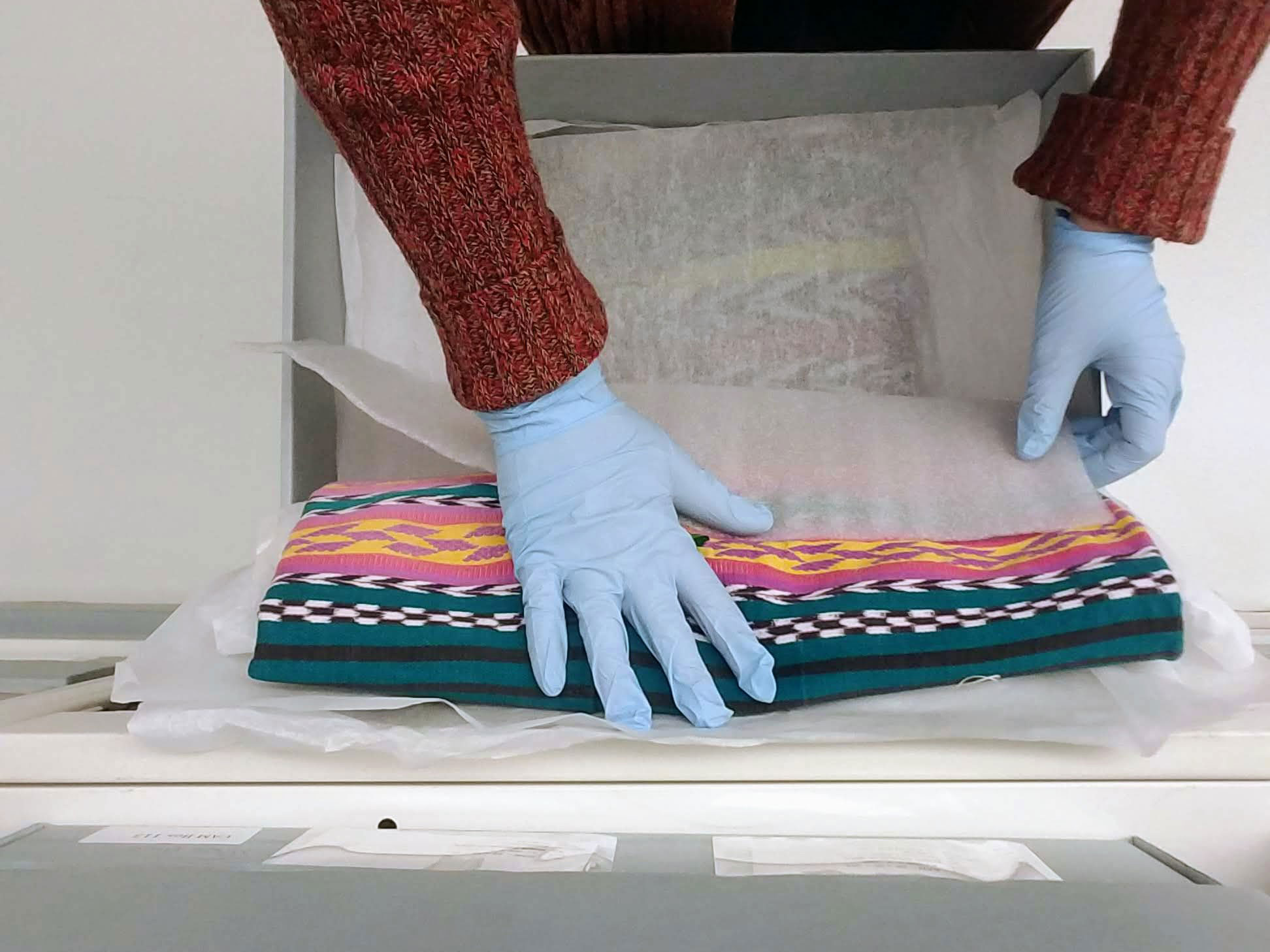 Gloved hands working with museum textile in drawer.