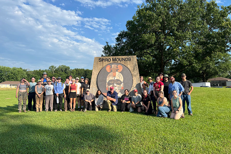 Students and faculty posing for a photo at the Archaeological Field School with a blue sky, trees, and grass.