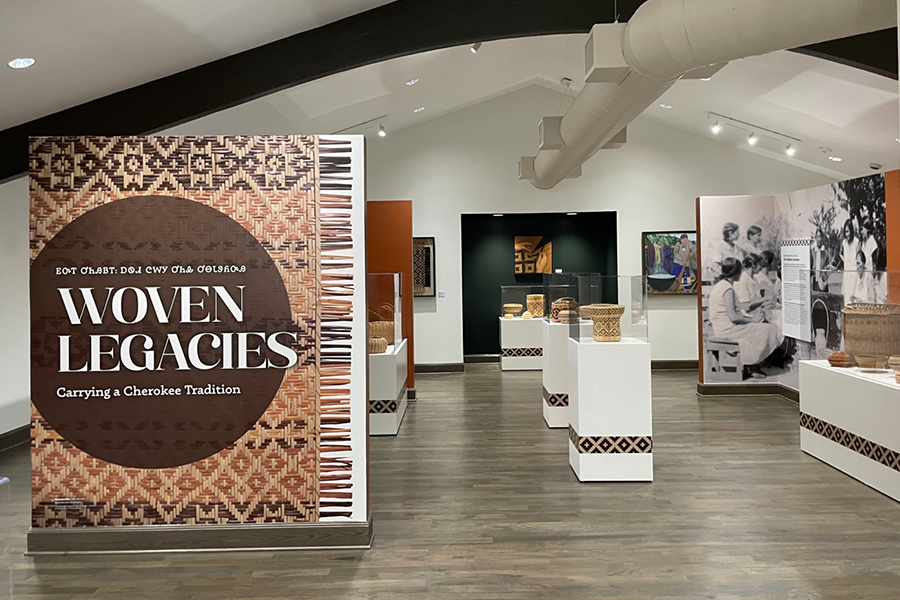 The Woven Legacies exhibit Evan Feely helped coordinate. In the left foreground, there is a large sign that reads "Woven Legacies: Carrying a Cherokee Tradition" with text written in Cherokee above. There are multiple woven baskets on display in the background. 