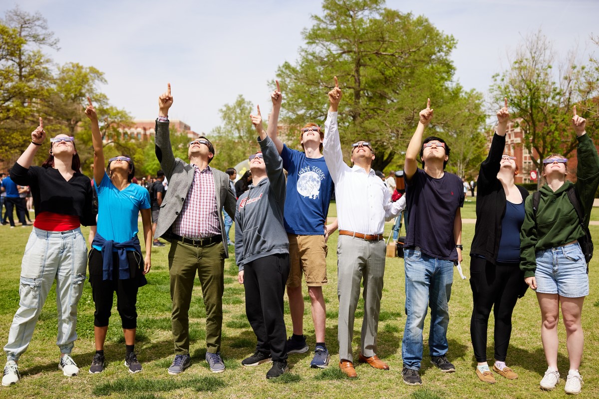 Attendees at the solar eclipse event stand side-by-side and point upward toward the sky while wearing solar eclipse glasses.