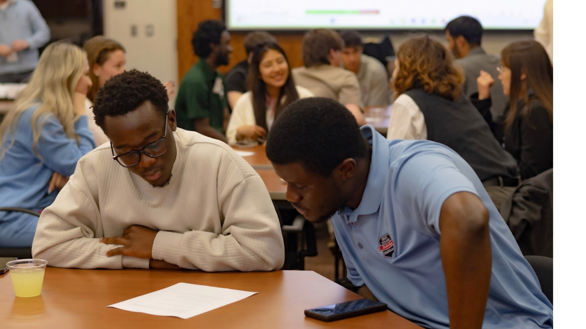 Two students around a round table with students in the background talking and smiling.