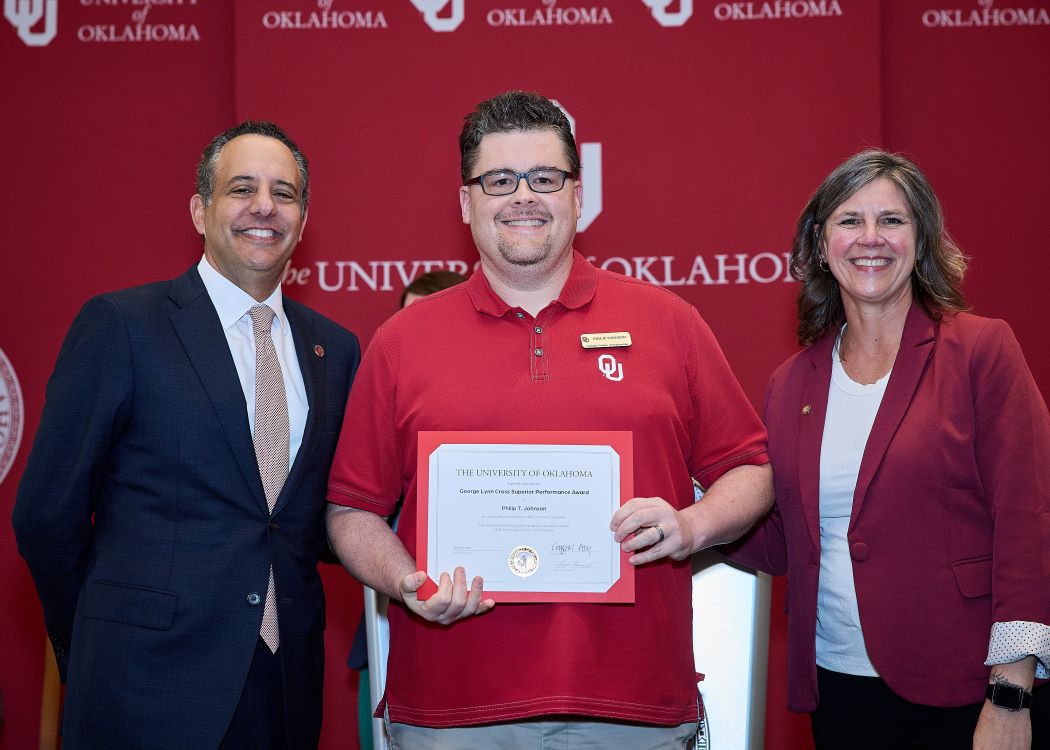 Philip T. Johnson holds an award certificate, standing with the University of Oklahoma president and a colleague in front of an OU backdrop