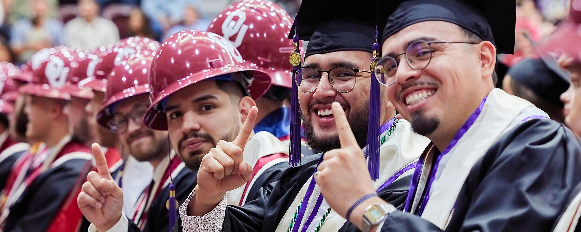 A group of graduates wearing regalia and raising their pointer finger during a ceremony.