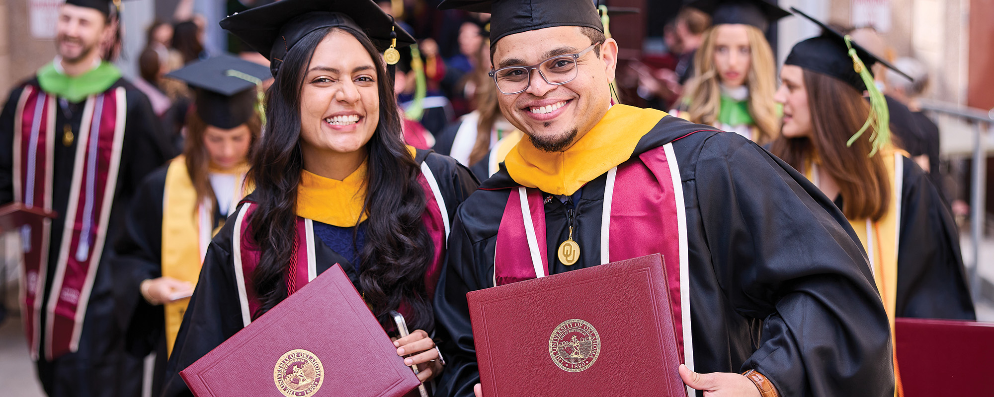 Two students in graduation regalia holding diploma covers and smiling at graduation.