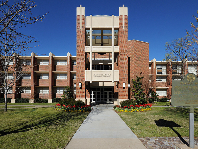 entrance of a red brick building with a tree on each side of a bell tower and grass in front