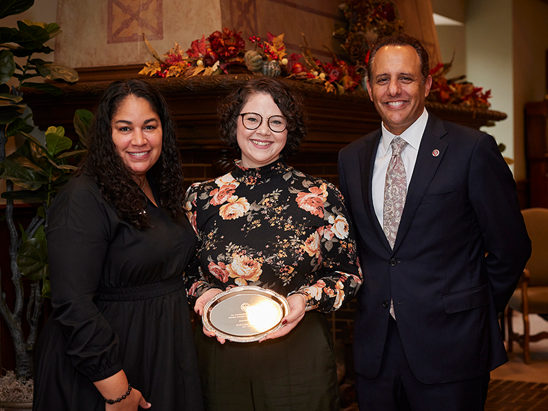A woman in black, a woman in a flower top holding a plaque and a man in a suit smile and post for a picture