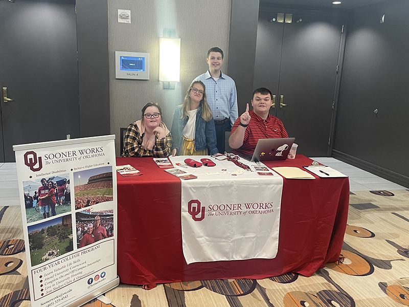 four students standing behind a table with OU swag in the lobby of a convention center