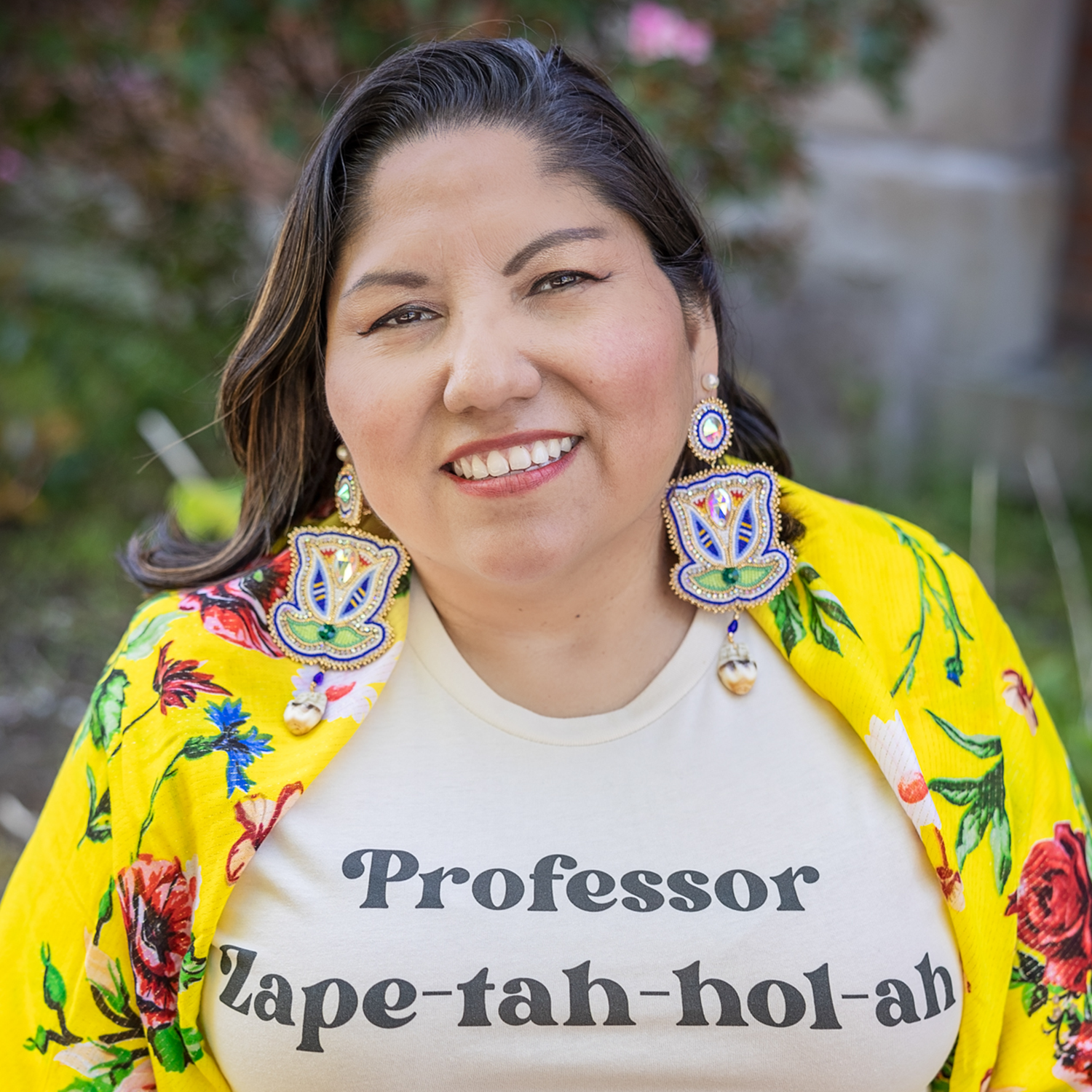 woman in bright yellow top, decoratve native earings wearing a shirt that says Professor Zape-tah-hol-ah