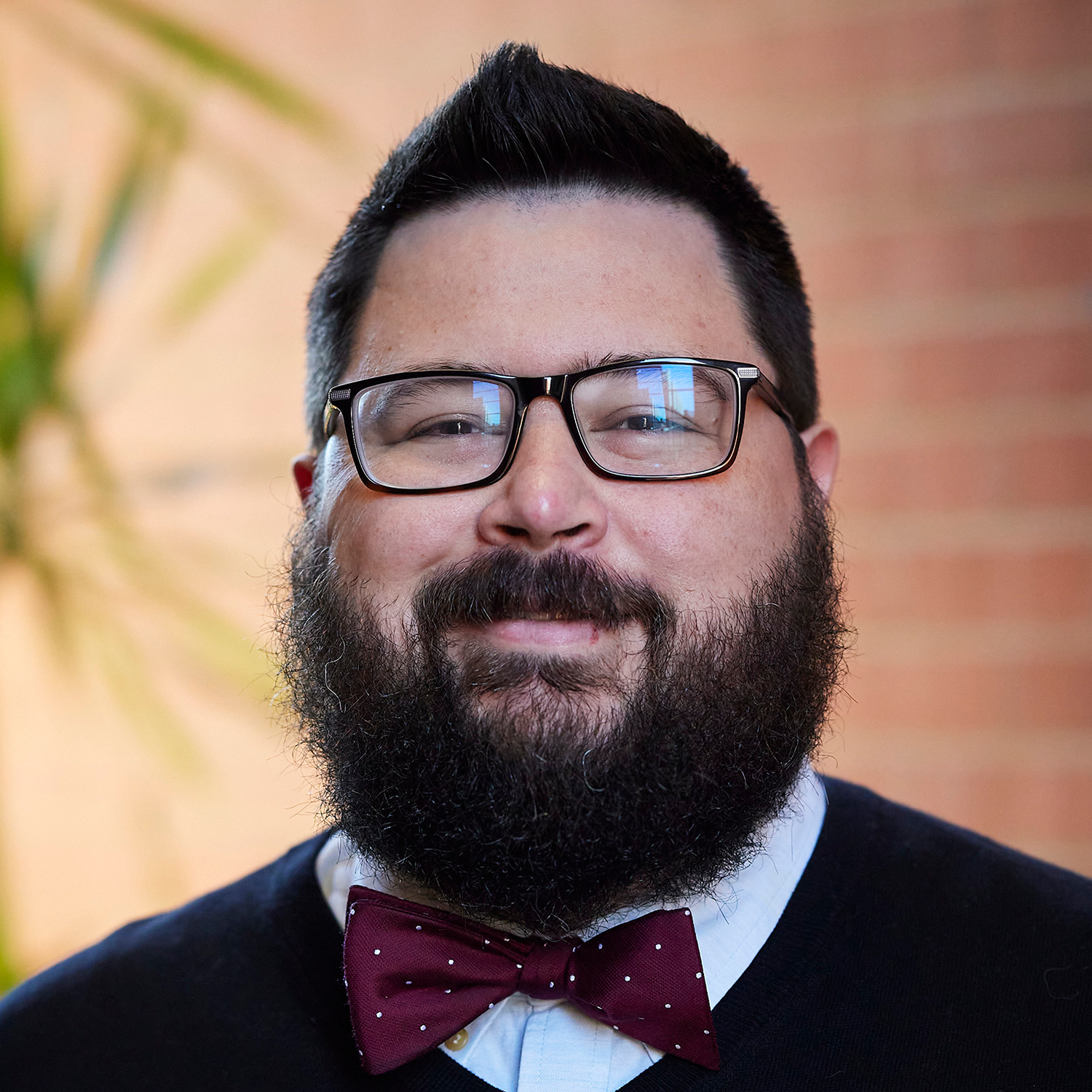 man in black sweater vest with red bow tie, dark hair and beard; glasses
