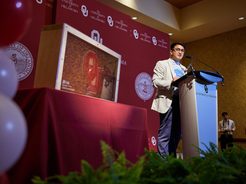 Man standing at podium speaking with a crimson OU backdrop
