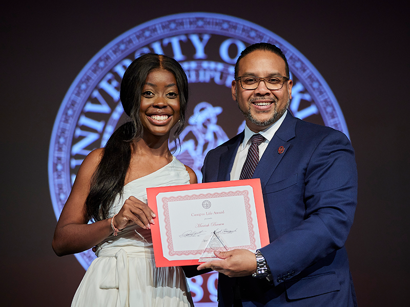 A woman on the left and a man on the right standing in front of the OU seal and holding an award certificate