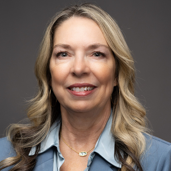 Woman in gray blue suit jacket and blue blouse, smiling at camera