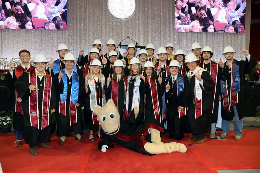 Gibbs College graduates in regalia and hard hats, with the Boomer mascot in the front of the group.