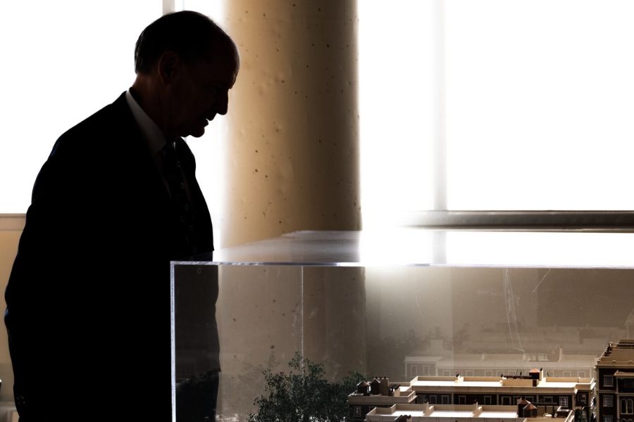 A man fully shadowed and facial expressions not visible looks over an architectural model that is encased in glass