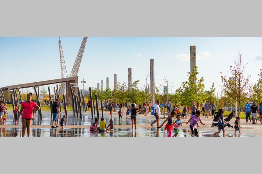 Children playing in splash pad with Butzer’s Scissortail Park shade pavilion and Skydance Bridge in the background.