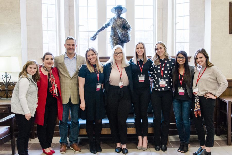 A group of professional looking people stand in front of the O U seed sower statue indoors