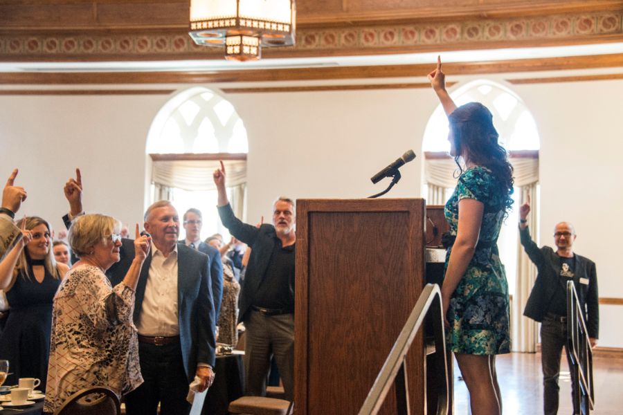 People standing up in ballroom with arms up and index fingers pointed up in OU salute.