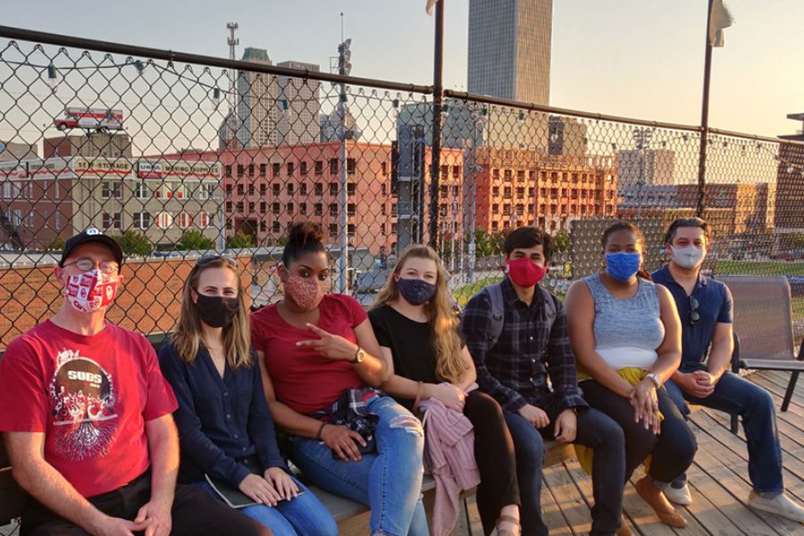 Eight masked students sitting on bench with city skyline behind fence