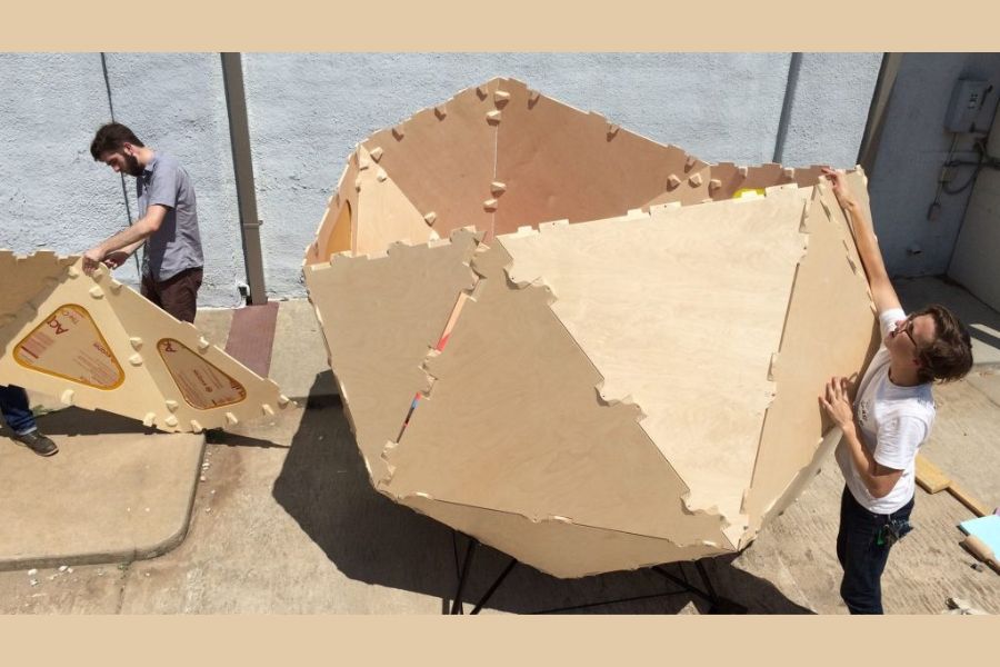 Students assemble a large geometric plywood structure outdoors using interlocking panels.