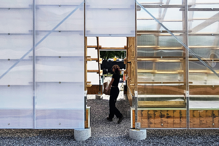 A person walks through the open central passage of a modular greenhouse constructed from translucent polycarbonate panels and exposed wood framing, with gravel underfoot and daylight filtering through the structure.