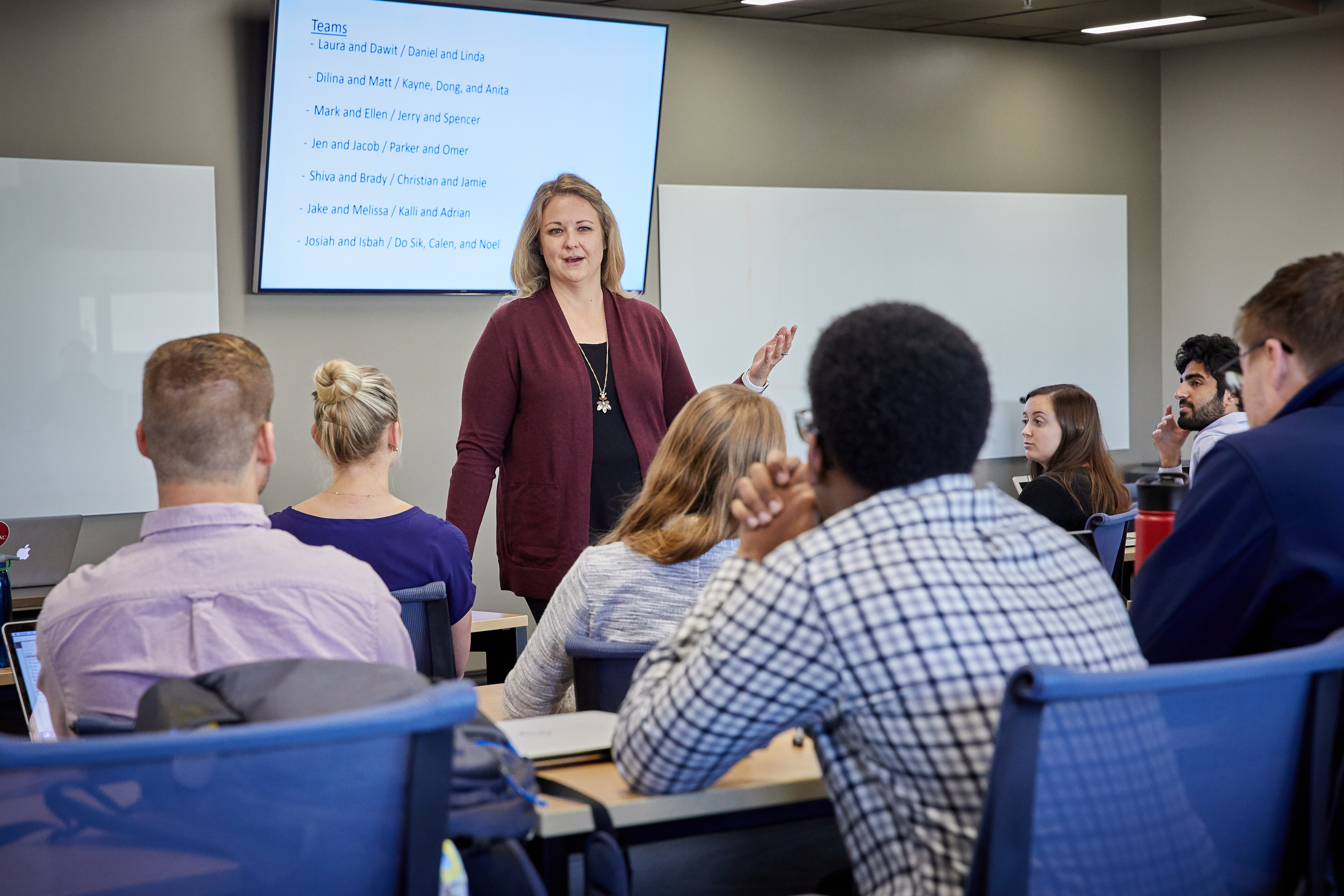 A faculty member stands at the front of a classroom, delivering a lecture to students who are seated and attentively listening. The professor is gesturing slightly, emphasizing key points while addressing the class. The students, facing forward, are engaged with the lecture, creating a focused and academic atmosphere.