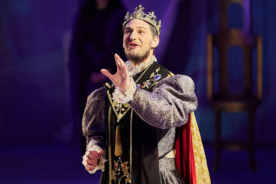 An actor in a crown, doublet and cape speaking with his hand raised.