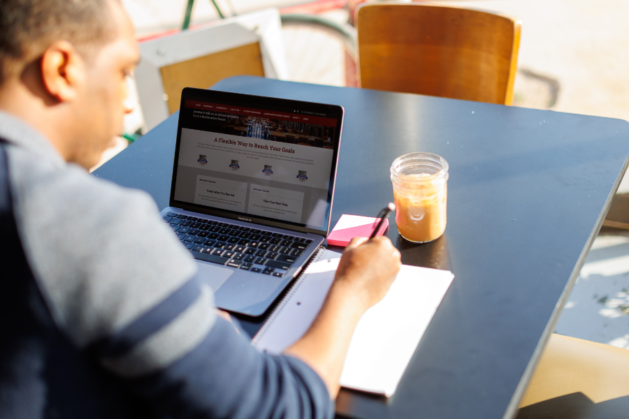 A student sitting at a table writes in a notebook while a laptop is open to a University of Oklahoma website that reads "A Flexible Way to Reach Your Goals."